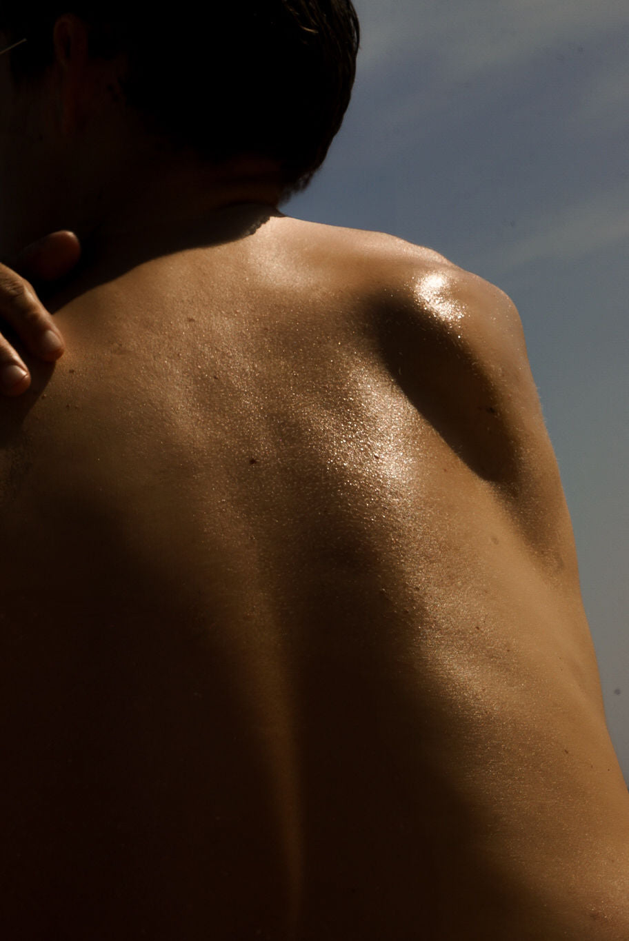 Sunlit close-up of a woman’s bare back and shoulder with glowing, dewy skin against a soft sky background.